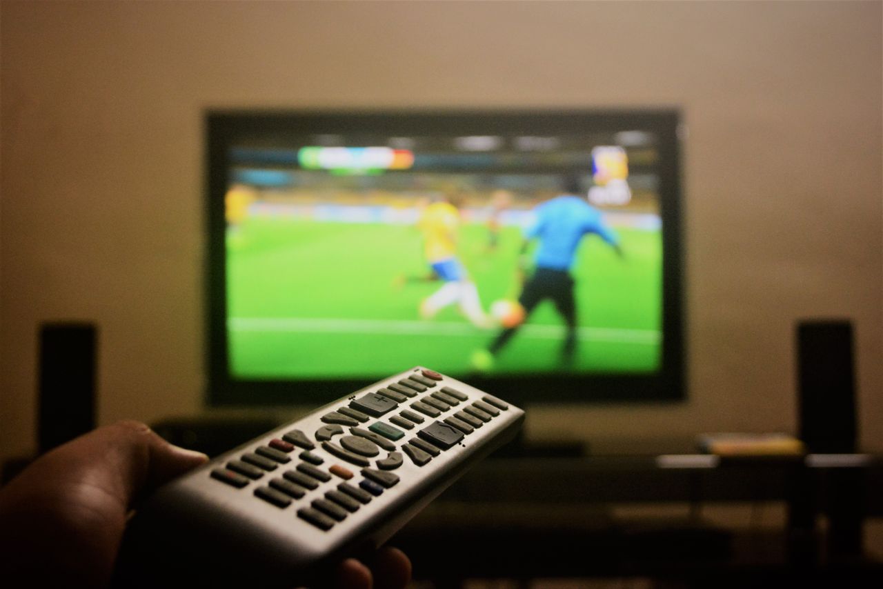 gallery-6 Close-up of a hand holding a remote control in front of a TV showing a football match.