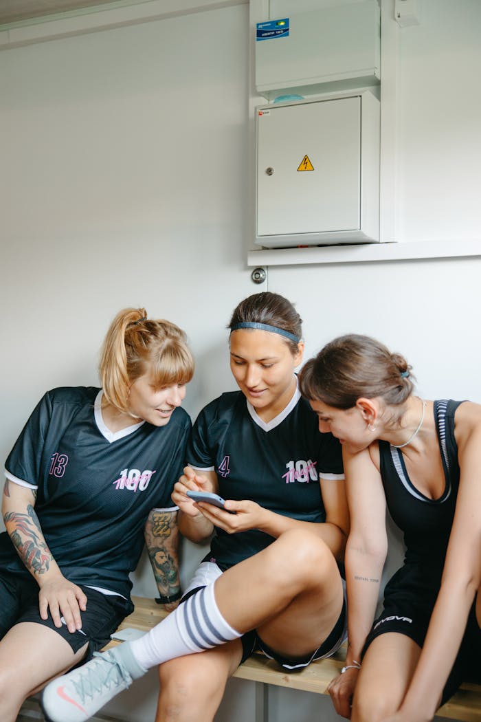 gallery-5 Three sportswomen in sportswear engage with a smartphone, sitting indoor on a bench.